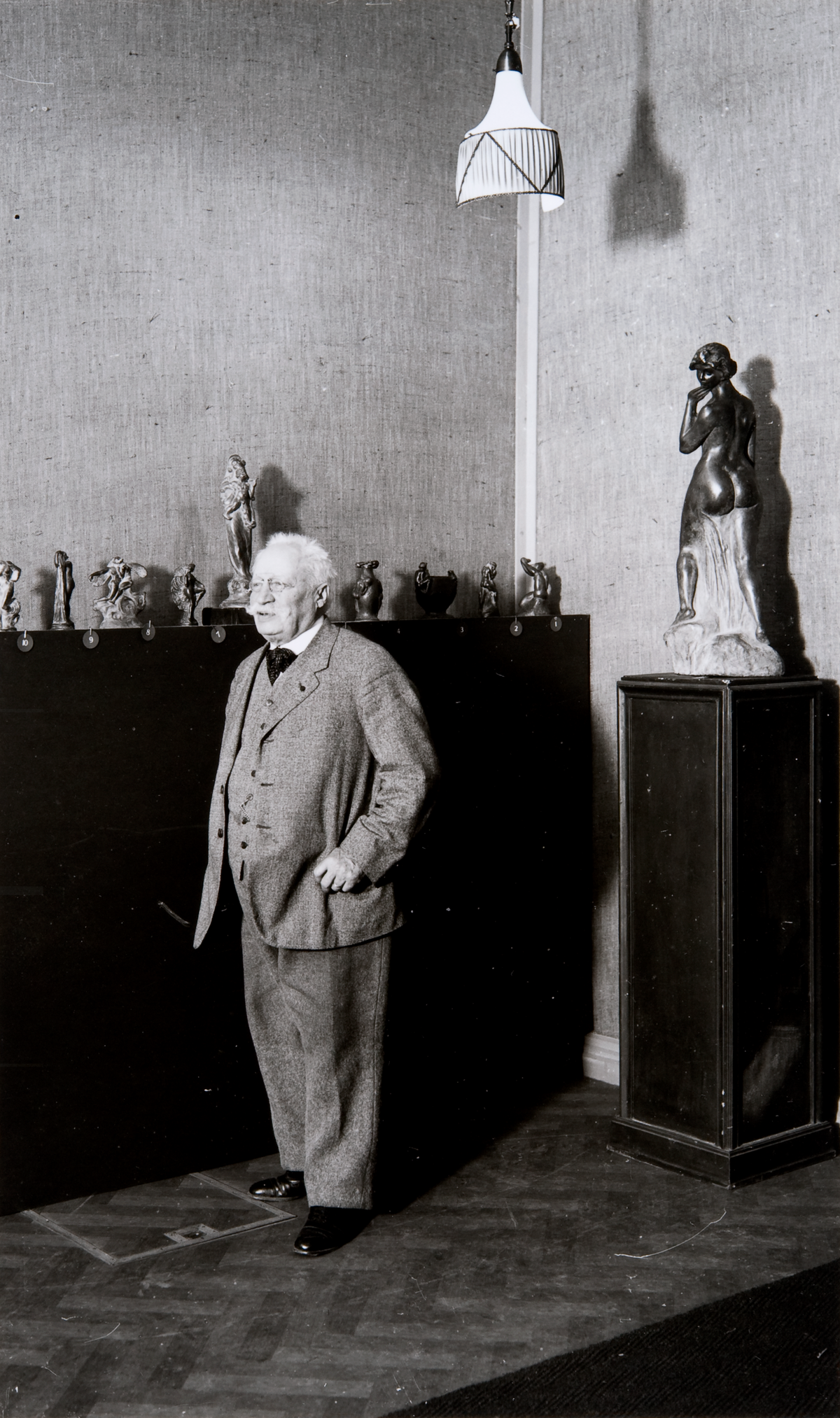 A black-and-white photo with a grey-haired gentleman with a gorgeous moustache standing in the middle. Small sculptures can be seen behind him, and a miniature version of Havis Amanda (a sculpture of a naked young woman) stands separate from the other sculptures.