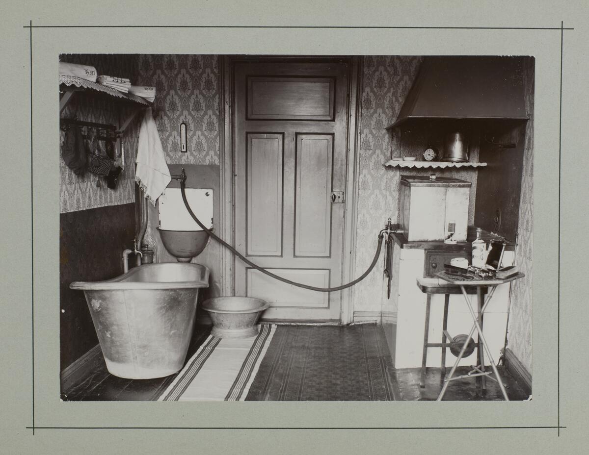 Kuvassa vasemmalla suuri kylpyamme, keskellä ovi ja oikealla puuhellan päällä seisova vesivaraaja, josta letku takaseinällä olevaan pesualtaaseen. Lattialla on matto ja seinillä kuviolliset tapetit.