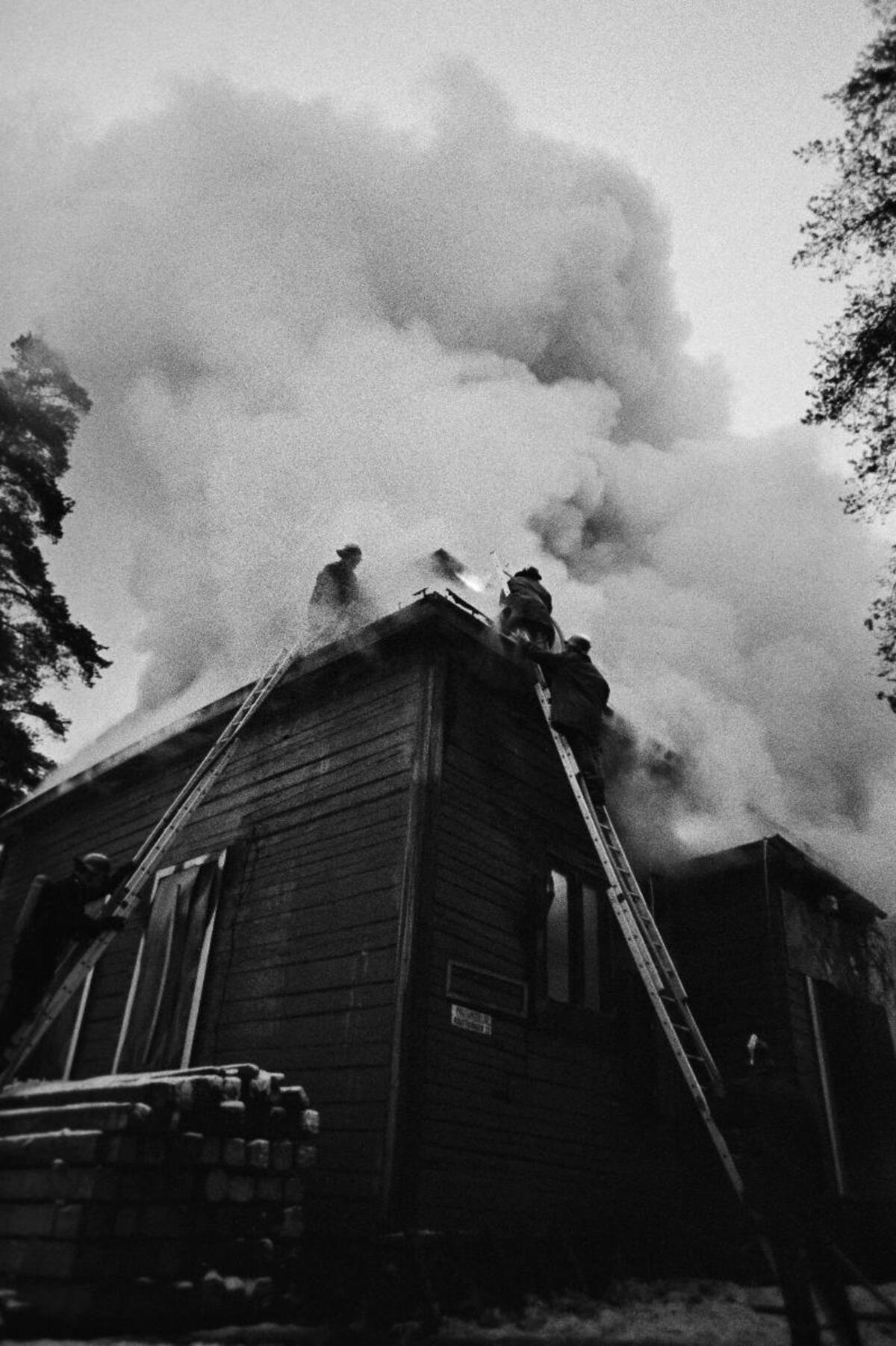 En brinnande stockbyggnad fotograferad nerifrån. Tre brandmän i släckningsarbeten. 