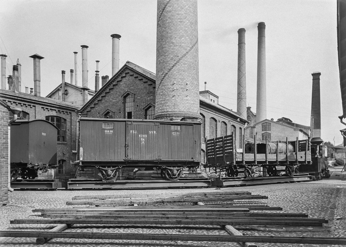 Etualalla maassa lautoja, kuvan keskellä junan tavaravaunuja ja niiden takana punatiilisiä tehdasrakennuksia savupiippuineen.