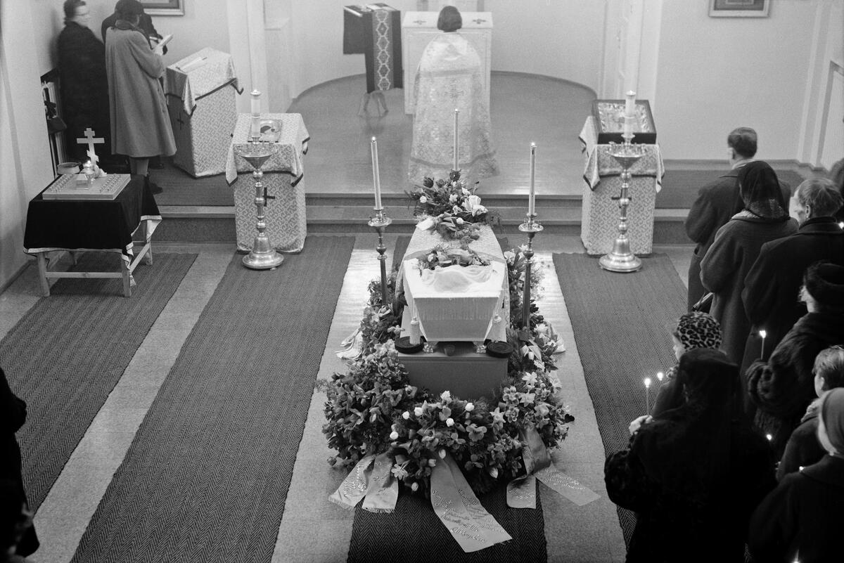 I mitten av bilden syns en likkista som omges av blommor. Bakom kistan står en präst med ansiktet mot altaret och ryggen mot kameran. På högra sidan av kistan syns begravningsdeltagare stående med ryggen mot kameran. De håller brinnande böneljus i handen. 