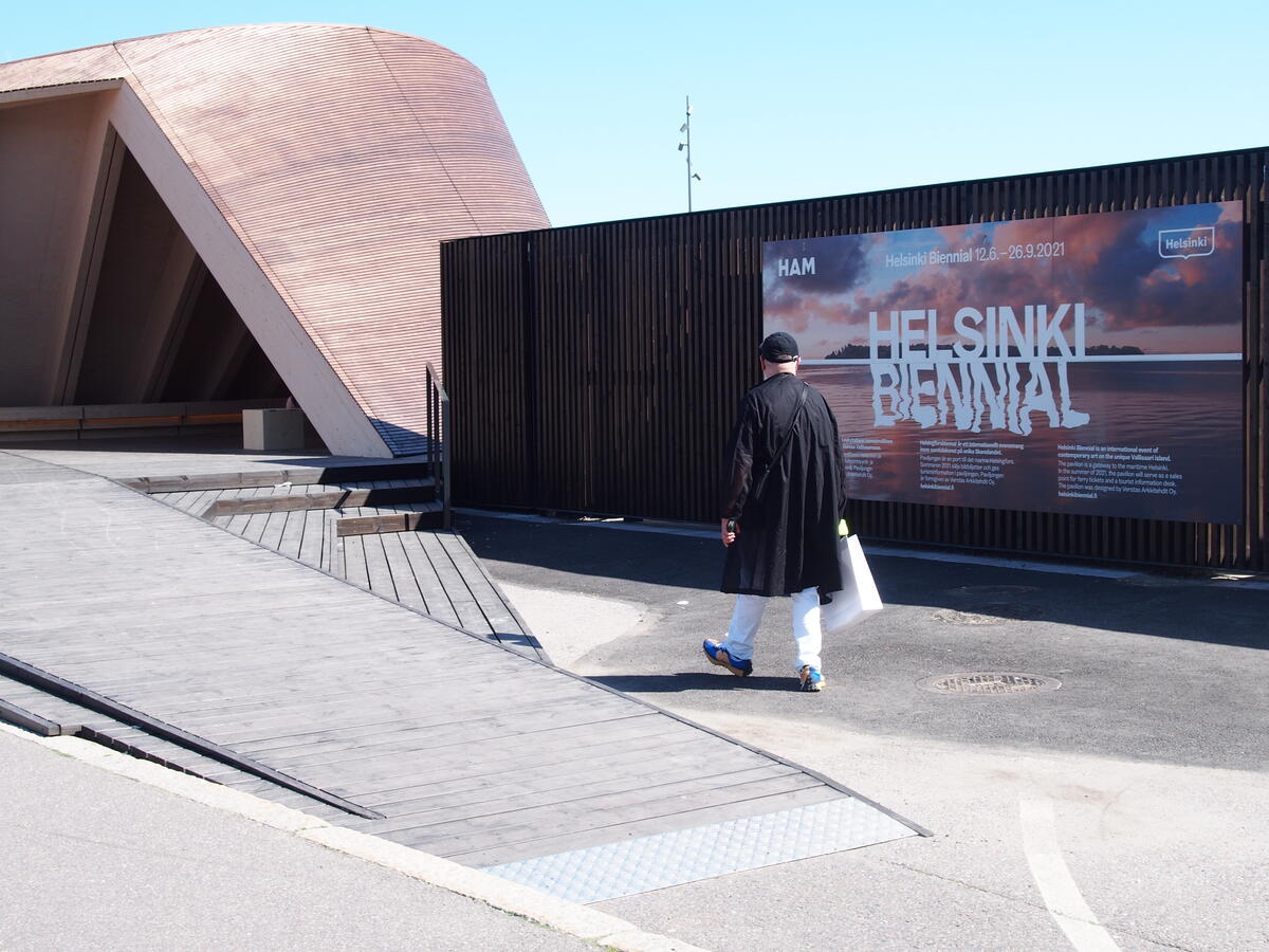 Helsinki Biennaalin mainos Etelärannassa. Etualalla takaapäin kuvattu, kävelevä ihminen.