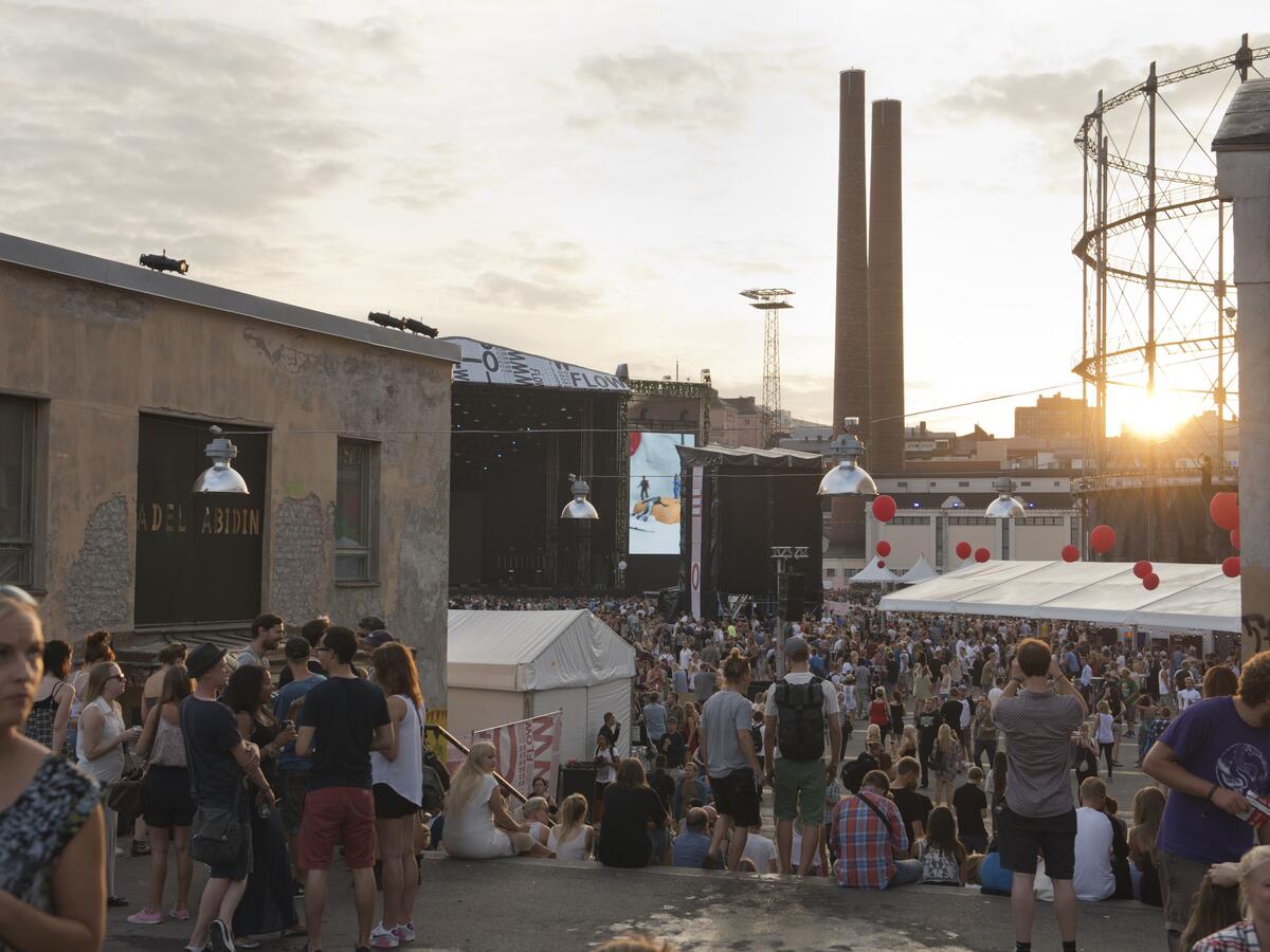 Flow-festivaalin yleisöä Suvilahden kesäillassa. Taustalla voimalaitoksen savupiiput ja kaasukellon teräskehikon taakse laskeva aurinko.