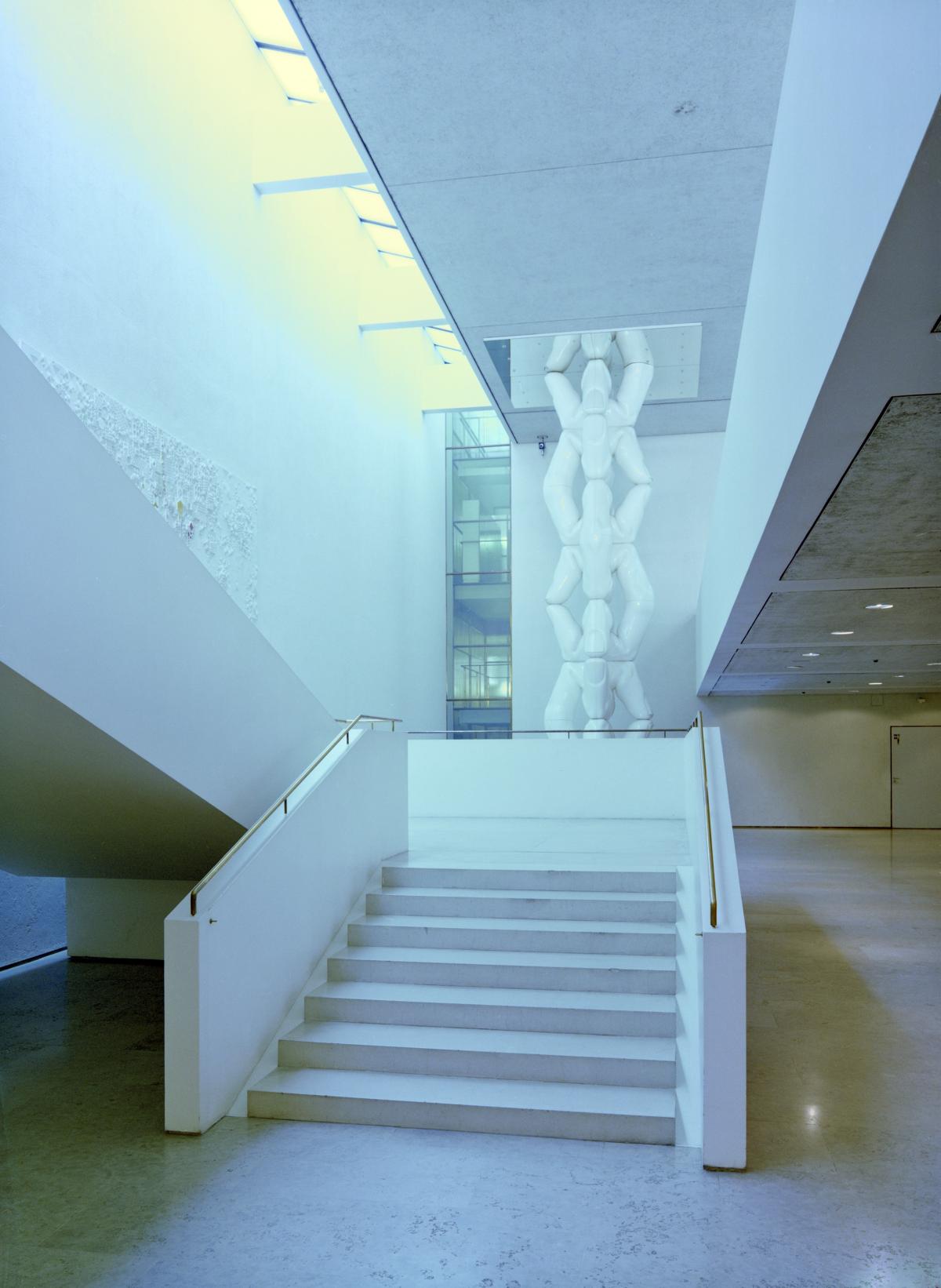 Vaalea portaikko Helsingin kaupungintalon aulassa. Luonnonvaloa laskeutuu portaisiin välikaton ja seinän raosta. Portaiden takana on vaalea veistostaideteos.