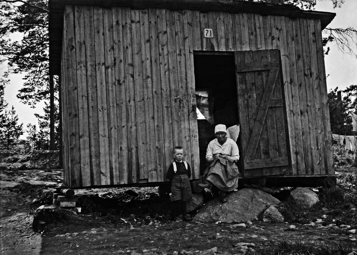 En kvinna och ung pojke framför ett av bräder snabbt ihopsatt tillfälligt hem som ursprungligen varit redskapsskjul, nära Tölö i Helsingfors. Kåken är byggd på en klippig backe.