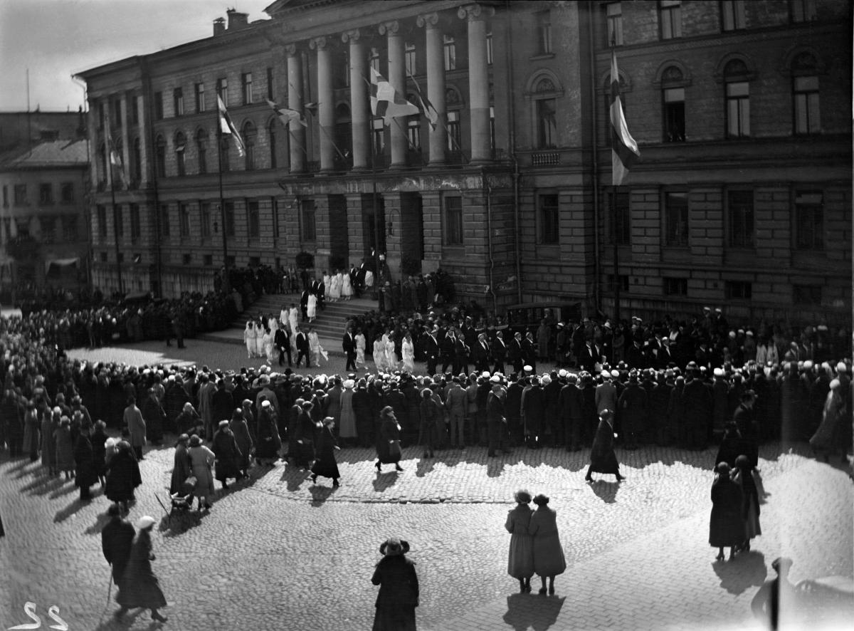 A long line of people walking in front of the University of Helsinki main building wearing black and white academic dresses