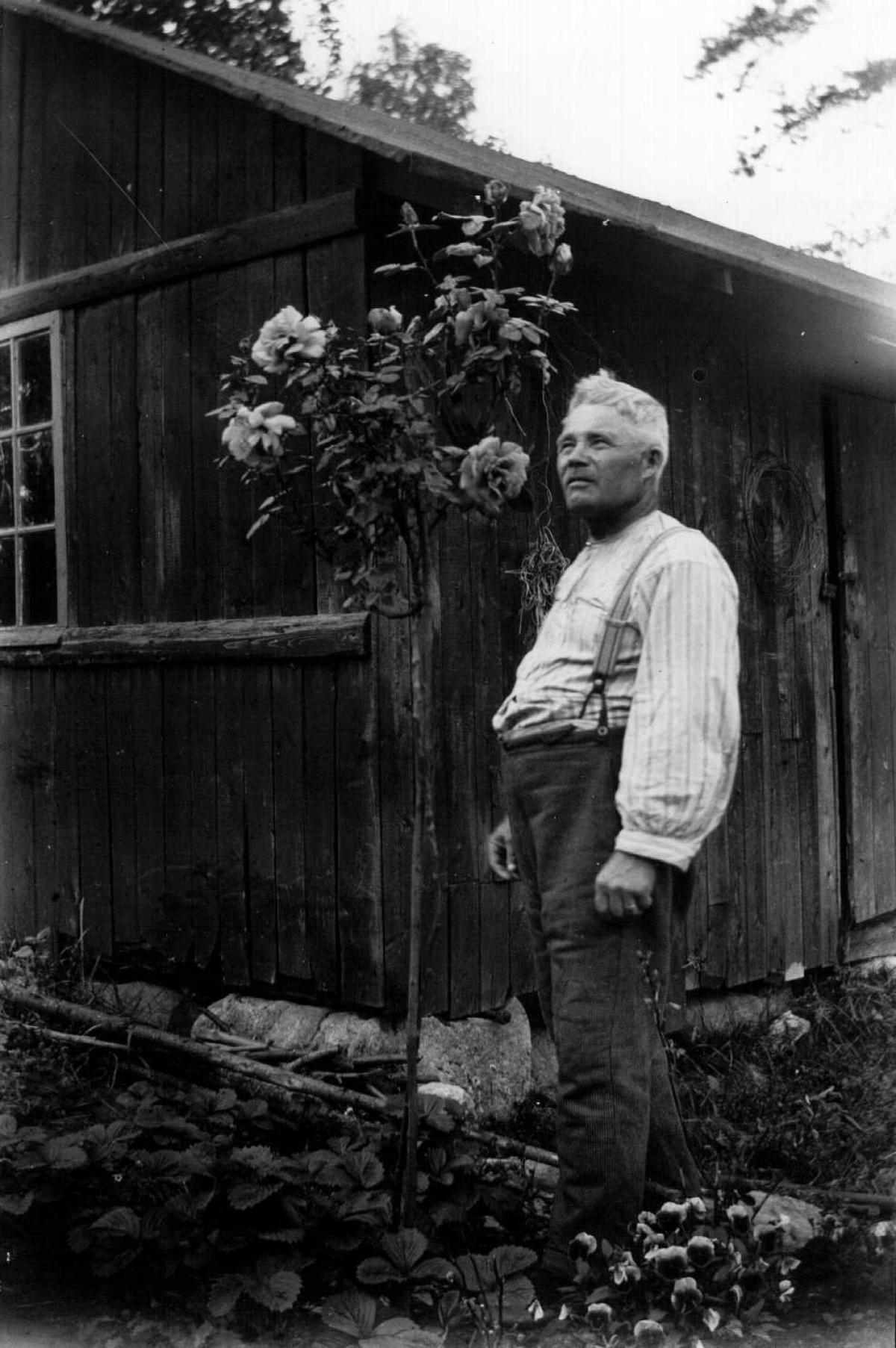 Vankkarakenteinen mies seisoo matalan puisen kotitalonsa edustalla. Hänen vieressään talon seinustalla on suuri kimppu kukkia.