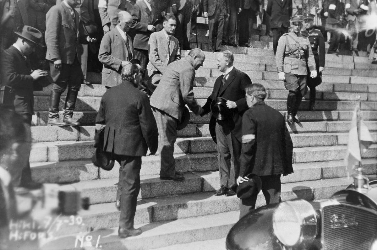 Two men shake hands on the stairs of the Cathedral. Several men around, some wearing white armbands.