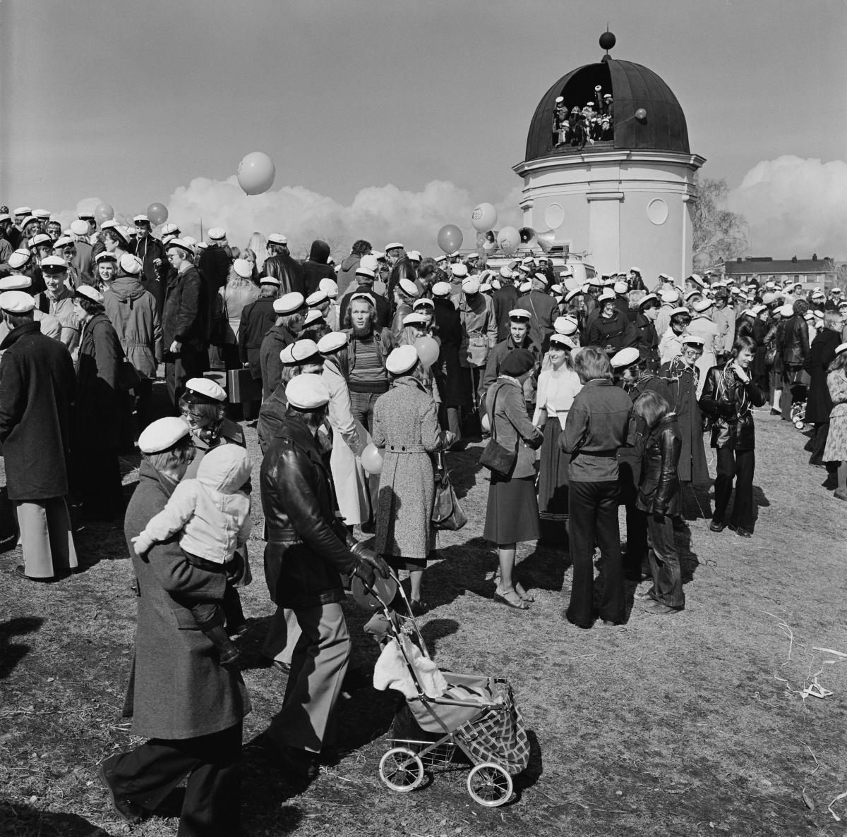 En stor skara människor klädda i studentmössor har samlats för att fira i Brunnsparken framför observatoriet. Observatoriets lucka står öppen och det finns festfolk även i kupolen. 