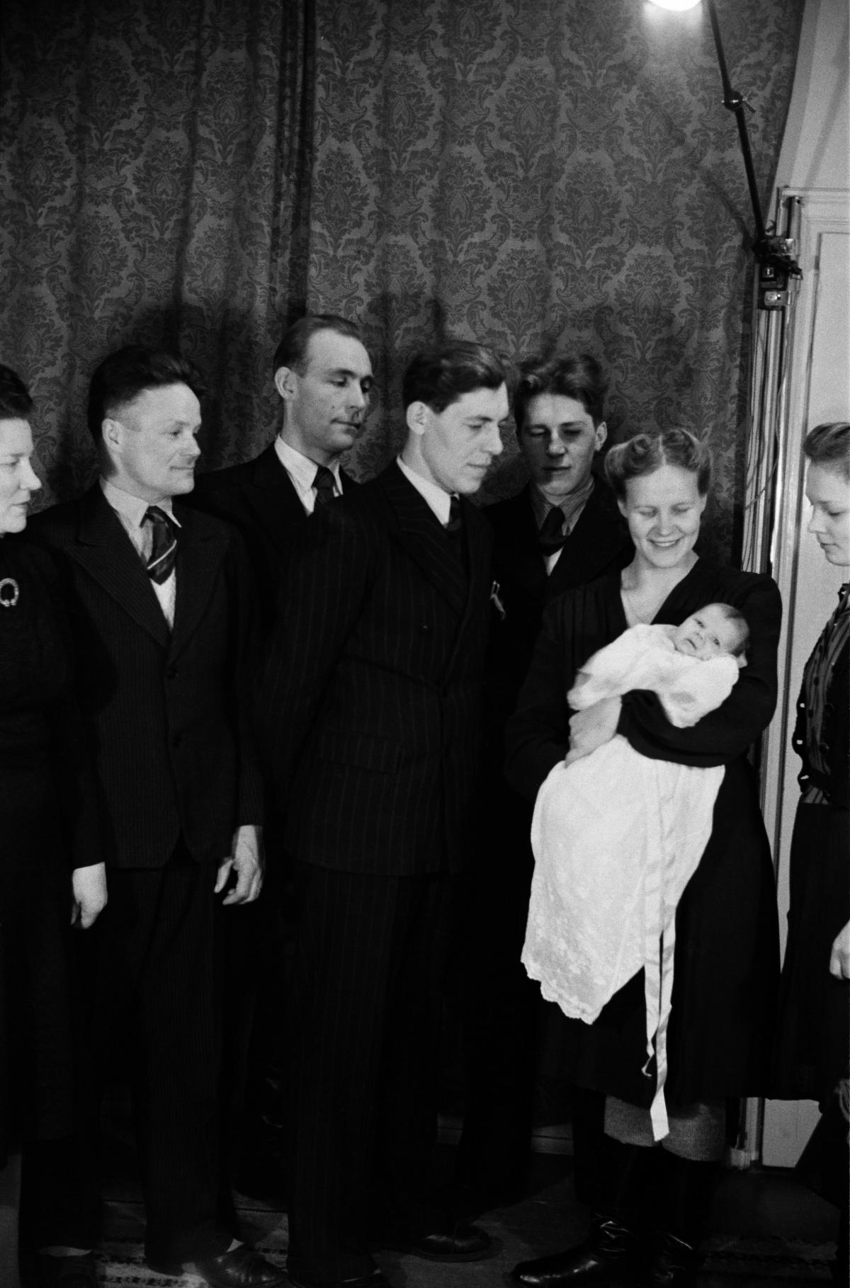 En leende kvinna håller ett barn i vitt dopklänning. Fyra män i kostym står bredvid och tittar på barnet. I bakgrunden fördragna gardiner med stor mönster.