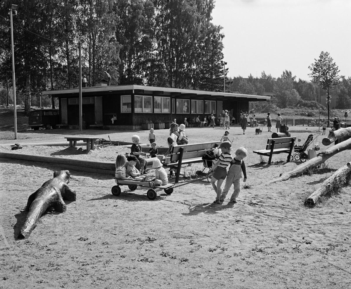 Gårdsplan vid Botbyhöjdens lekplats. Två småpojkar drar en kärra med andra barn ombord. På gården finns en sandlåda, bänkar, stockar och en krokodil av trä.