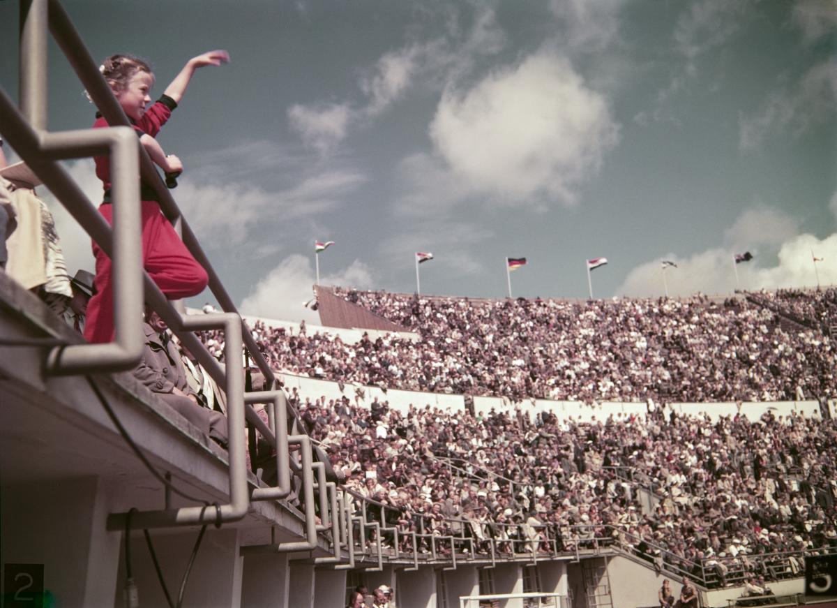 A wide shot of the northern stand of the Helsinki Olympic stadium, which is full of spectators. On the front, a young girl in a red dress waves her hand.