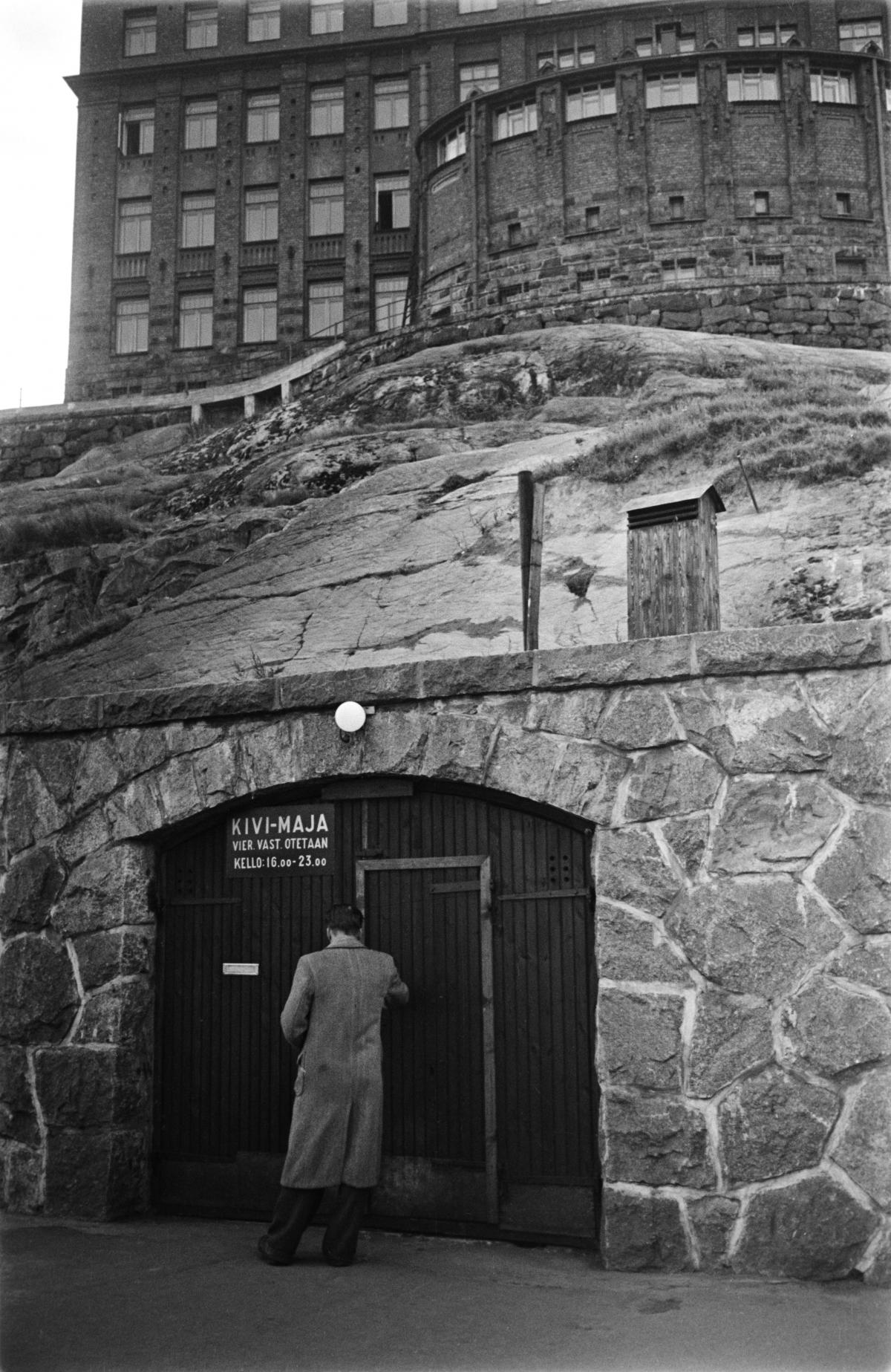 Mies seisoo asunnottomien yömaja Kivi-Majan oven edustalla Siltavuorenrannassa. Yömaja toimi vanhassa väestönsuojassa ja sisäänkäynti on kallioseinämässä. Taustalla näkyy yliopiston Siltavuorenpenkereen rakennuksia.