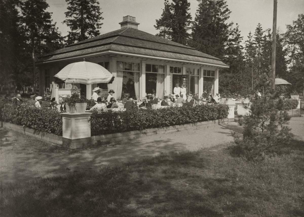 Ett svartvitt foto som föreställer en vacker gammal kafébyggnad. På terrassen sitter kvinnor i ljusa klänningar och hattar.
