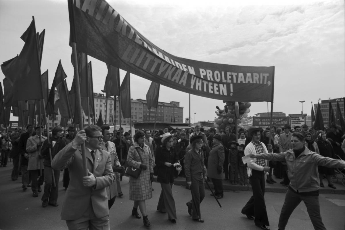 Työväen vappumarssi lähdössä liikkeelle Hakaniemestä. Yleisö seuraa kulkuetta, jossa kannetaan punaisia lippuja. Eturivissä kannetaan banderollia jossa teksti "Kaikkien maiden proletaarit liittykää yhteen!"