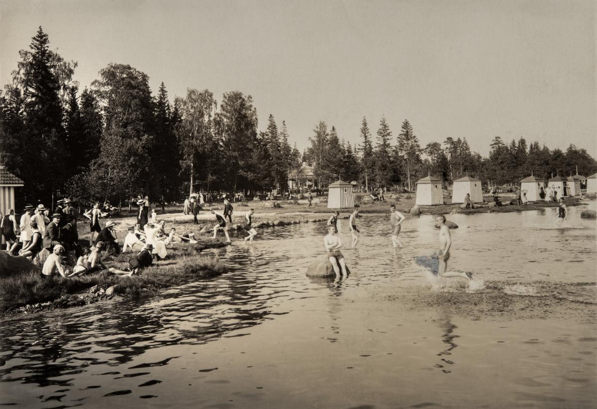 Människor simmar på simstranden. I bakgrunden syns tältliknande badhytter. 
