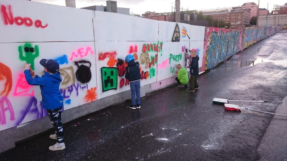 Nuoria lapsia maalaamassa Suvilahden graffitiseinällä. Lapset ovat piirtäneet nimiään ja yksinkertaisia hahmoja. Taustalla seinällä graffitiharrastajien monimutkaisempia teoksia.