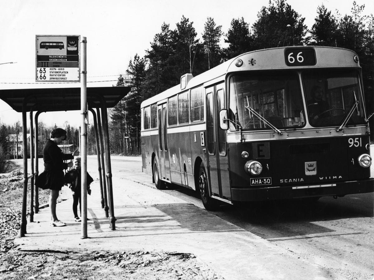 Buss nummer 66 vid hållplatsen, under hållplatstaket står en kvinna och ett barn.