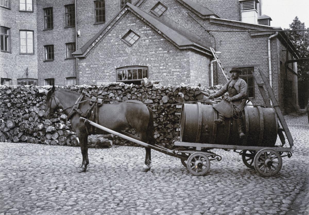 Ölkusk iklädd förkläde sitter på en bryggerivagn formad som en tunna med en häst spänd framför. 