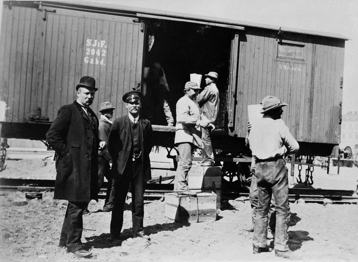 Arbetare lastar en öppen godsvagn på bangården till Helsingfors gamla järnvägsstation. Förutom arbetarna poserar en tågtjänsteman i sin uniform samt en man klädd i snygg kostym och hatt. 