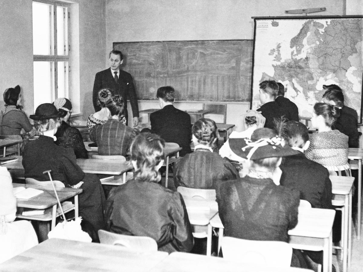 Lektion i historia på de äldstes dag. Litet klassrum fyllt med elever klädda enligt festen. Läraren står framför klassen, bredvid honom en krittavla och en stor karta över Europa. 