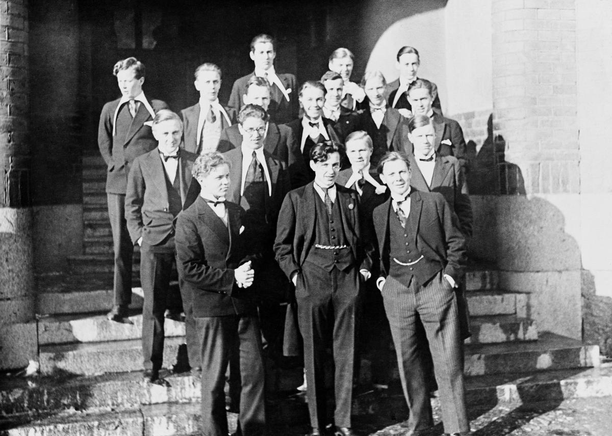 En grupp pojkstuderande på trappan till finska normallyceum. Pojkarna är stiligt klädda i sina fars kostymer. En del bär vita skjortor med mycket långa hängande kragar.