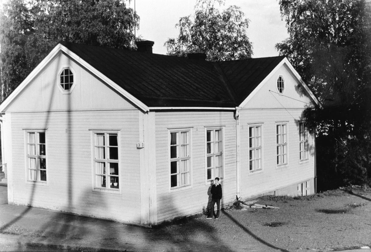 Vaalea puinen kaksikerroksinen harjakattoinen rakennus näkyy kuvassa etualalla. Talon seinustan vieressä seisoo nuori lyhythiuksinen ihminen. Rakennuksen ympärillä on puita.