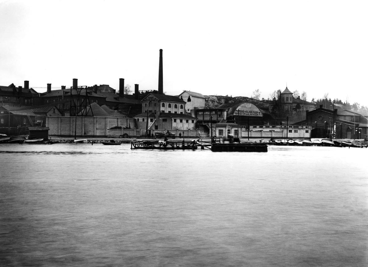 Murbruksfabrik på Sörnäs strandväg fotograferad från havet. På gaveln till den buktade byggnaden texten "PARGAS KALKSBERG AB / PARAISTEN KALKKIVUORI OY".