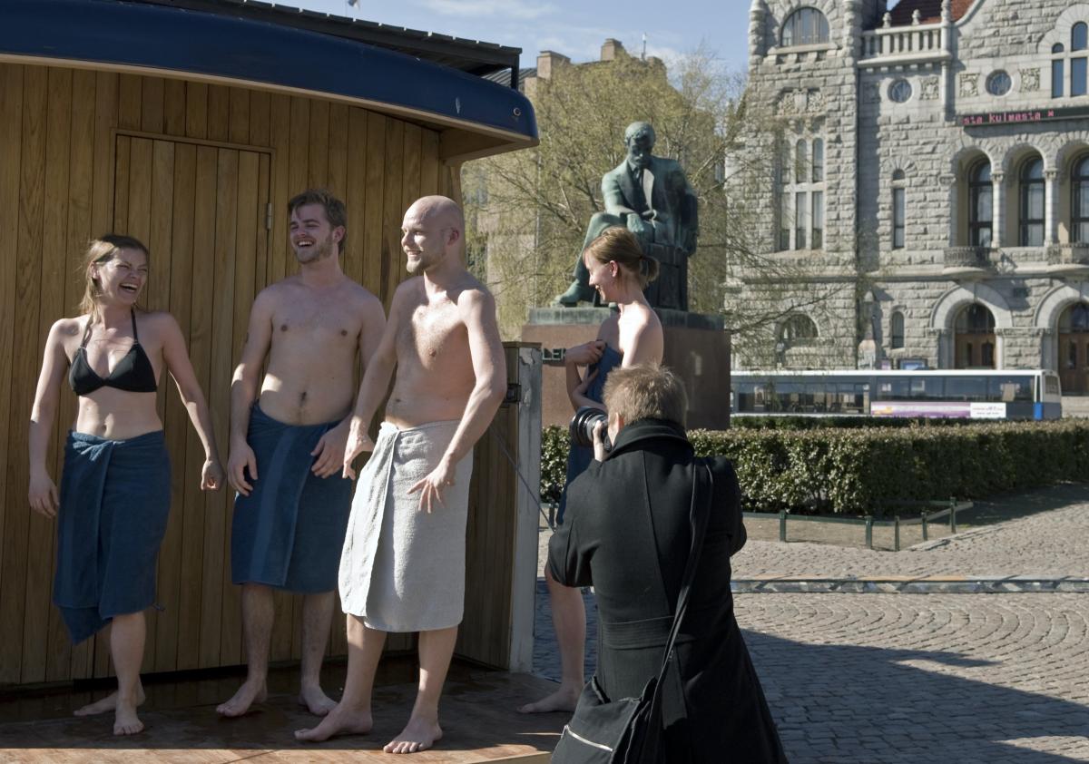 Kaksi miestä ja kaksi naista pyyhkeisiin verhoutuneina siirrettävän saunan edessä. Taka-alalla Aleksis Kiven patsas ja Kansallisteatteri, etualalla selin valokuvaaja.