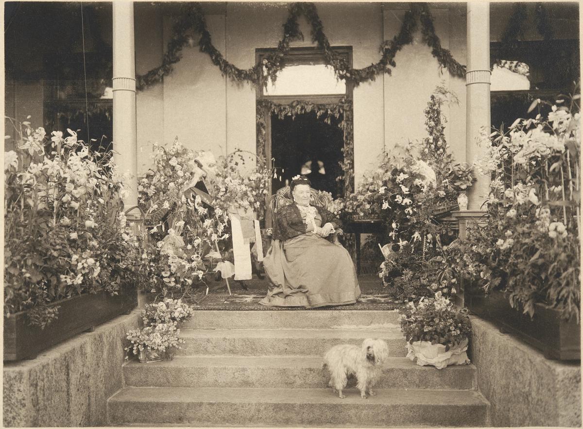 Aurora Karamzin fotograferad på sin 90-årsdag sittande framför ytterdörren till sin villa, omringad av blomplanteringar. En liten ljus hund sitter framför henne på trappan. 