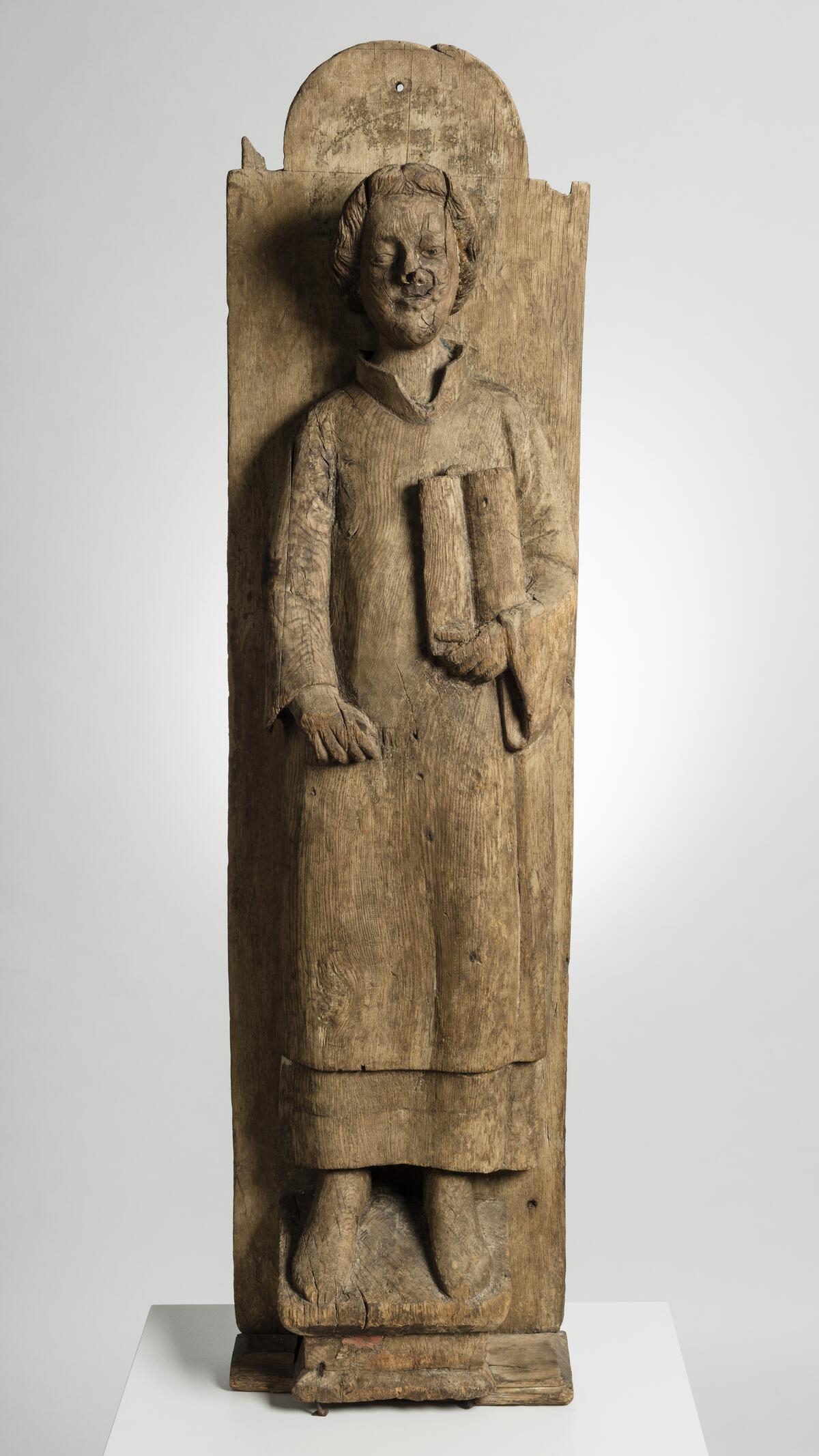 En omålad träskulptur som föreställer Sankt Laurentius. Han håller en öppen bok i ena handen. Skulpturen är fäst vid ett bräde.