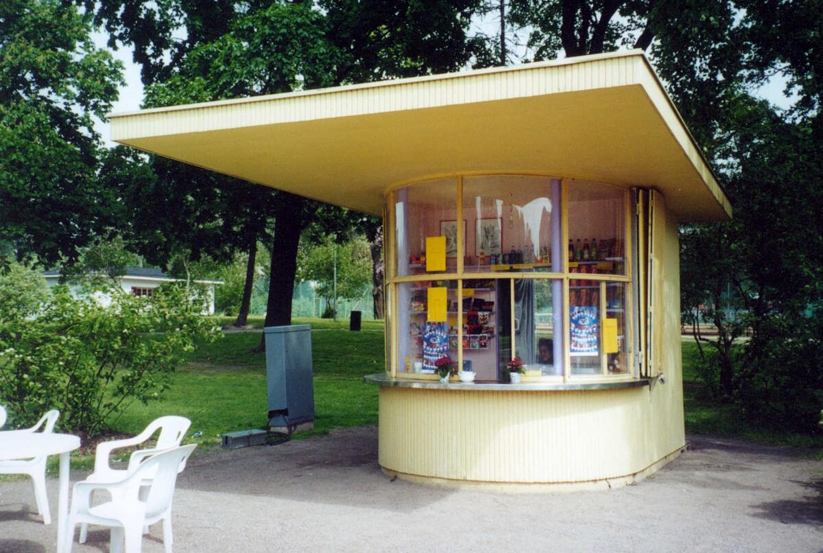 Ljusgul kioskbyggnad, där den främre delen är rundad och fönsterförsedd. Takskägget sticker ut långt framför kiosken.