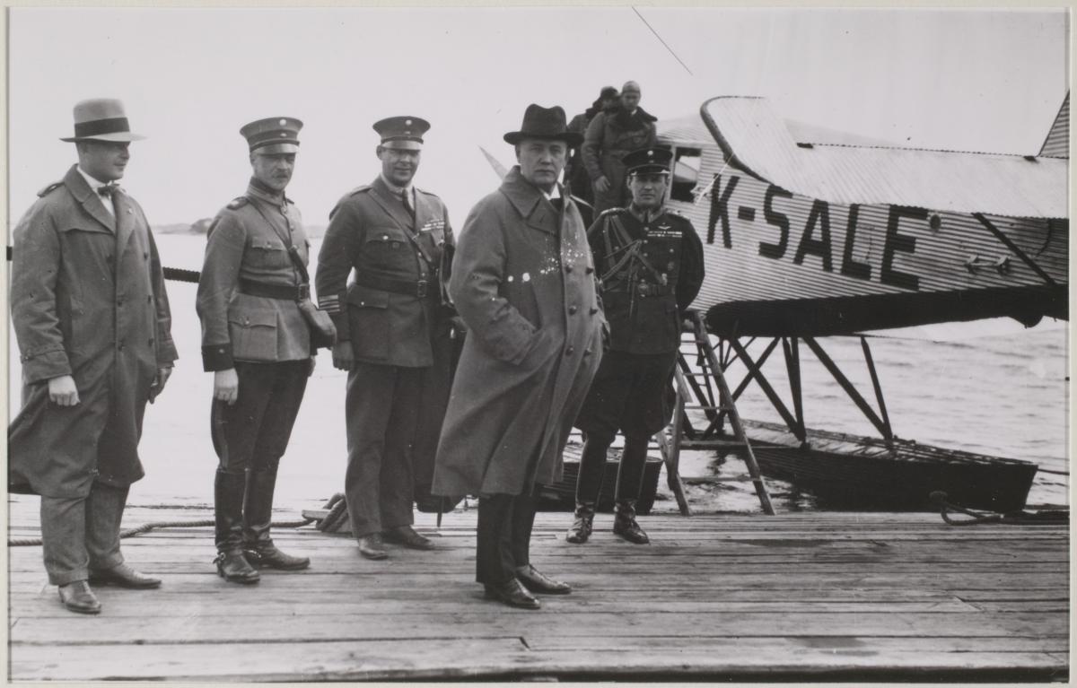 Päällystakkiin ja hattuun pukeutunut presidentti Relander seisoo lentosataman laiturilla. Presidentin takana vesitaso ja univormuasuisia adjuntantteja.