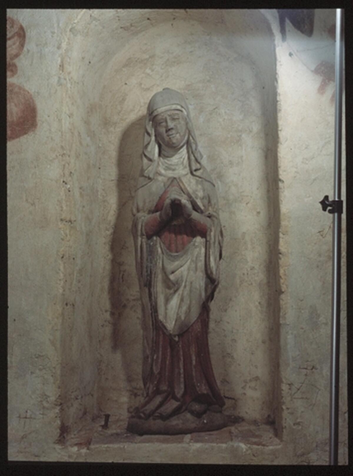 En stenvägg med nisch, i den står en träskulptur som föreställer en kvinna i huvudduk, lång klänning och kåpa. Hon håller sina händer höjda framför sig som i bön. 