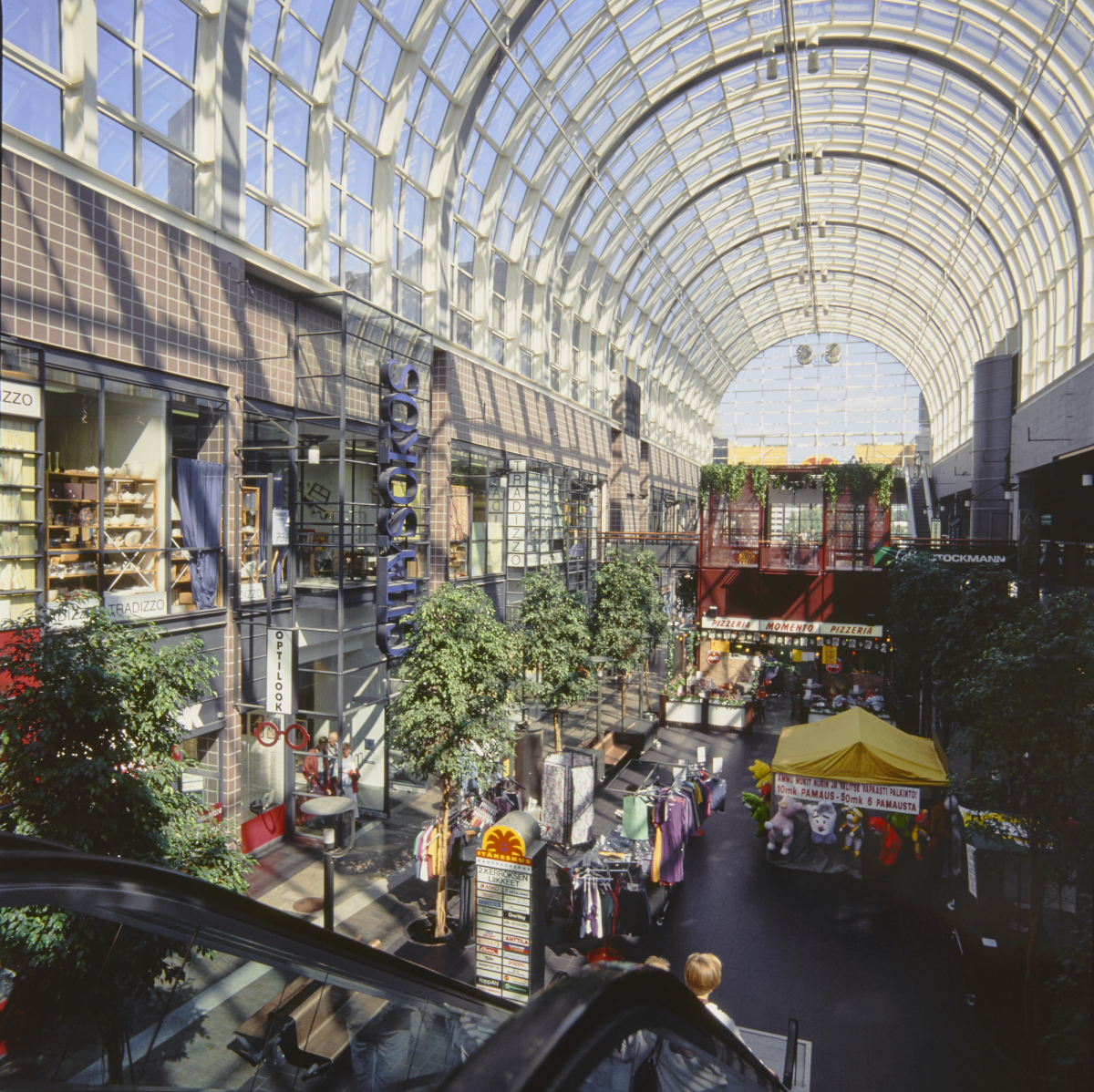 Itäkeskuksen kauppakeskus sisältä kuvattuna 1990-luvulla. Päivänvalo pääsee kauppakeskukseen kaarevan lasikaton läpi. Käytävällä on lukuisia koristepuita. Liiketilojen lisäksi käytävällä on myyntiteltta.