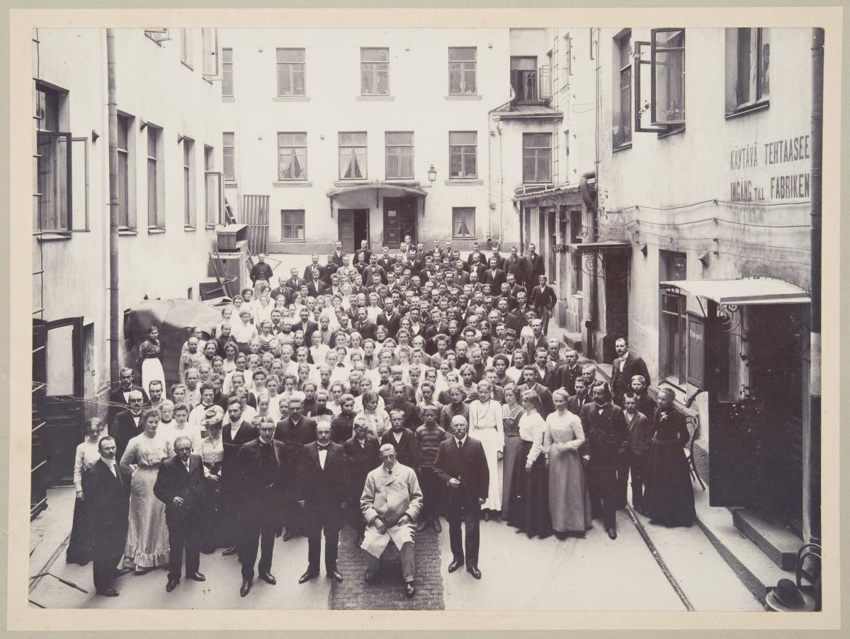 Gruppfoto av Weilin & Göös personal på innergården av sin arbetsplats. Tiotals människor har ställt sig på bilden. I förgrunden i mitten sitter Karl Gustav Göös klädd i ljust. 