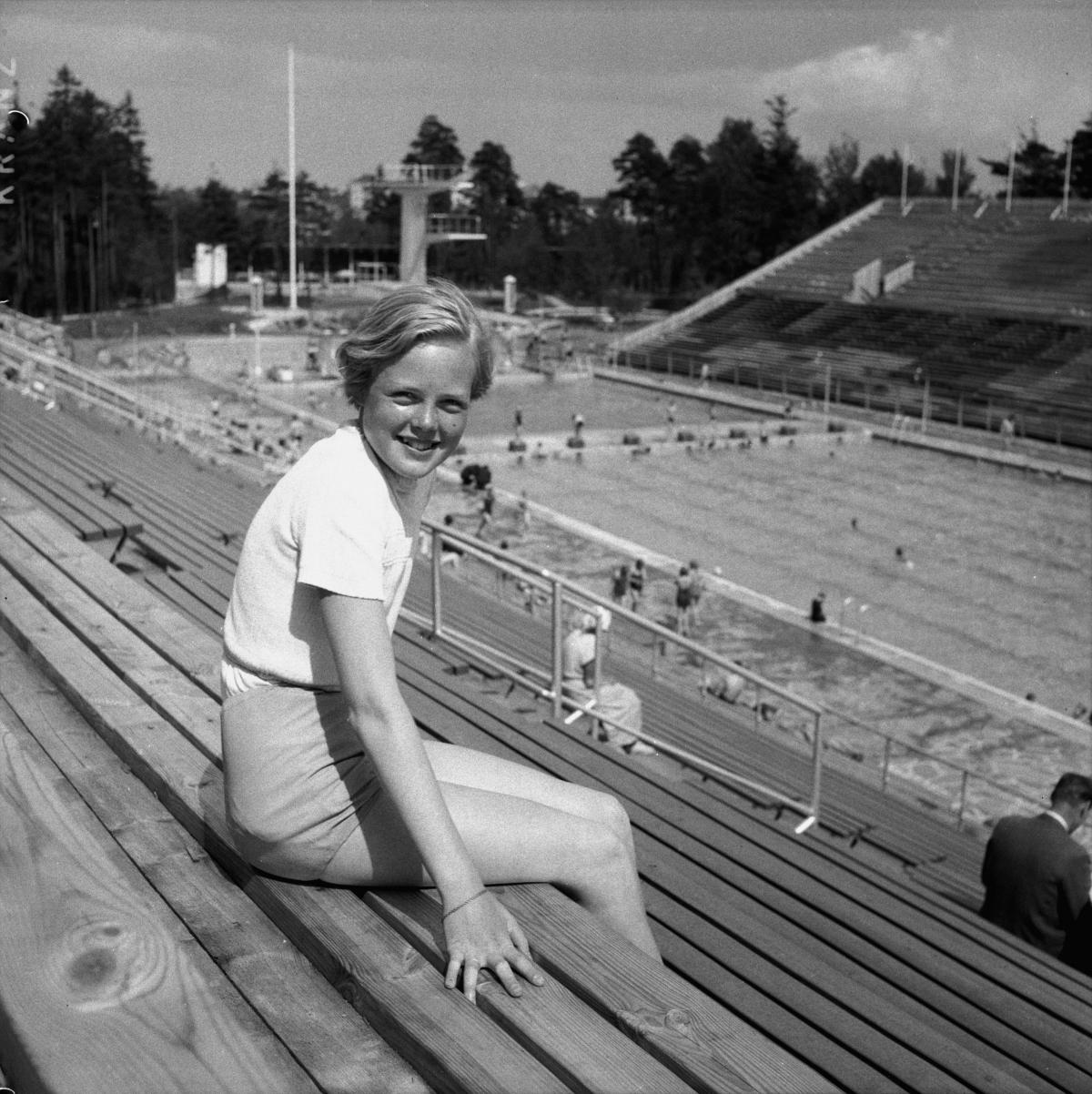 Nuori nainen istuu uimastadionin katsomossa kesällä 1952. Taustalla stadionin allas ja uimahyppytorni.