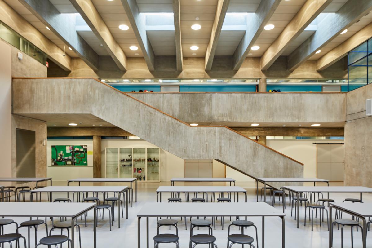Roihuvuoren ala-asteen keskussali, jossa pöytiä ja tuolia. Saliin laskeutuu raskas betoniportaikko koulun toisesta kerroksesta.