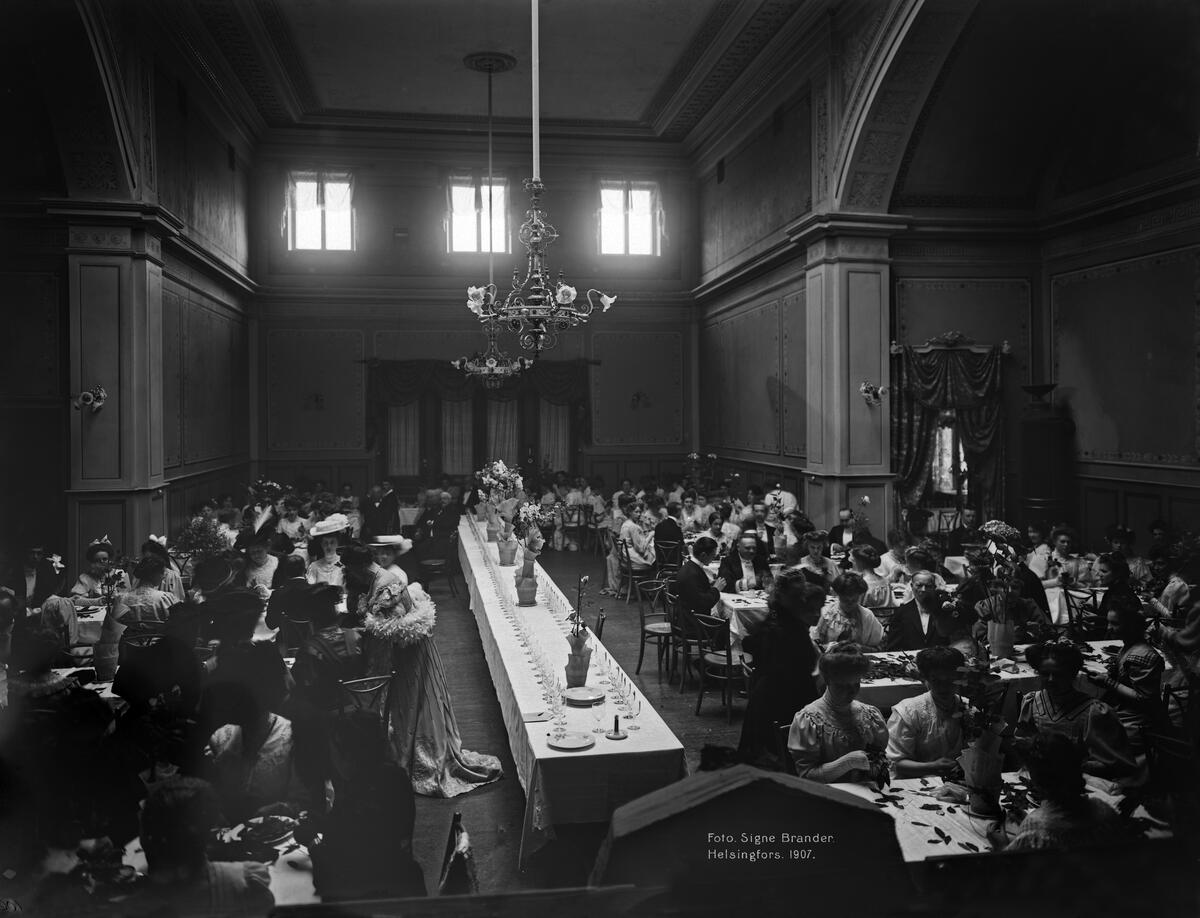 Naisia ja miehiä valmistamassa laakeriseppeleitä pöytien ääressä. Keskellä kuvaa pitkä pöytä, jonka päällä on kukkaistutuksia. Pöydän päällä suuria kattokruunuja.