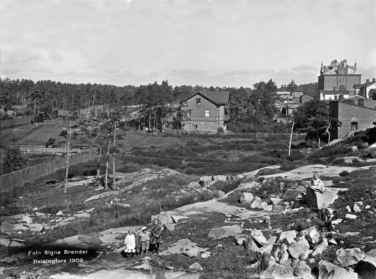 Mustavalkoinen kuva, jossa oikealla joitain rakennuksia, yhtenä Vallilan kansakoulu. Etualalla maisema on kallioista, taaempana puita. Aivan kuvan etuosassa seisoo kolme lasta. Oikeassa kulmassa istuu yksi lapsi kalliolla.