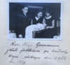Kastemekkoon puettu lapsi naisen sylissä. Ympärillä seisoo mies ja nainen, kaikki katsovat lapseen.
